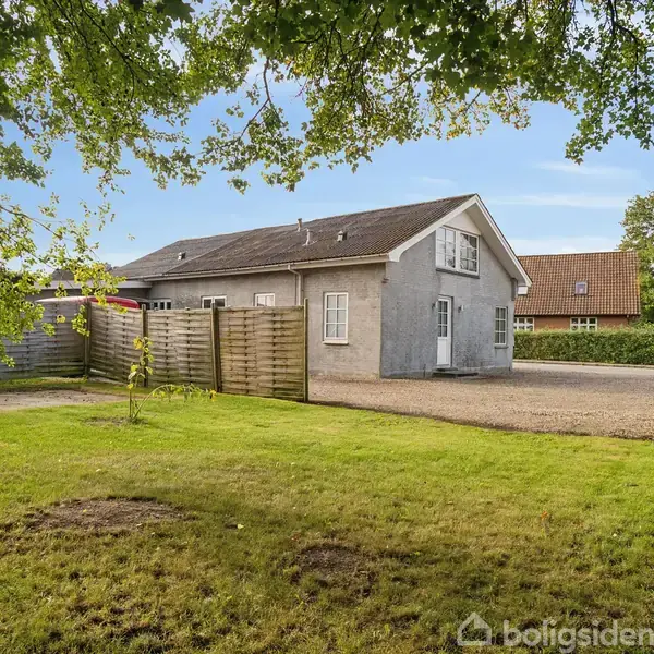 Grå bygning med skråt tag står stille i en åben gård, omgivet af grøn plæne og træer, ved siden af et plankeværk og grusområde.