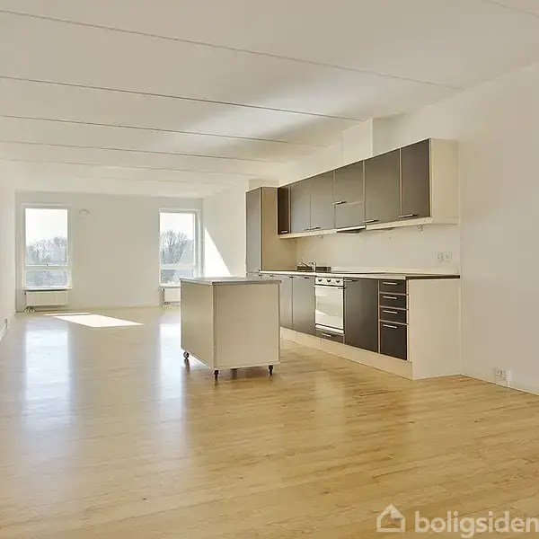 Køkken med gulv-til-loft vinduer i baggrunden, åbne skabe og skuffer langs væggen samt en flytbar ø i midten, i en lys, rummelig stue.