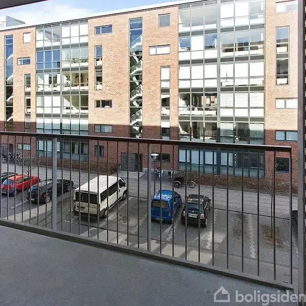 Balkon med metalrækværk ser ud mod en moderne bygning med murstens- og glasfacade. I gården nedenfor er parkerede biler og cykler, der giver en urban atmosfære.