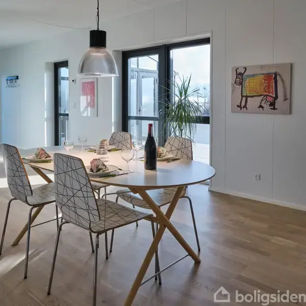 Et rundt spisebord med fire stole er dækket med service og en flaske vin. En pendellampe hænger over bordet. I baggrunden ses en plante og et maleri på væggen.