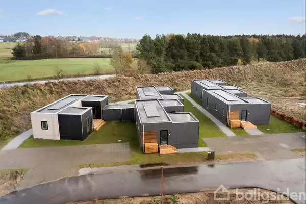 Modulære, moderne huse ligger stille i række med grønne tage og trappetrin foran. De er omkranset af græsarealer og en skov i baggrunden under en blå himmel.