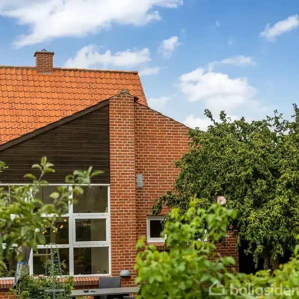 Rødt murstenshus med rød tegltag står stille omgivet af frodig grøn beplantning og træer. Store vinduer vender mod en blå, letskyet himmel.