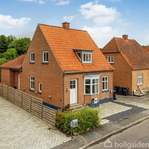 Et rødt murstenshus med skråt orange tegltag står på en stille villavej. Huset har en lille trappe ved hoveddøren og er omgivet af en flisebelagt indkørsel og hæk.