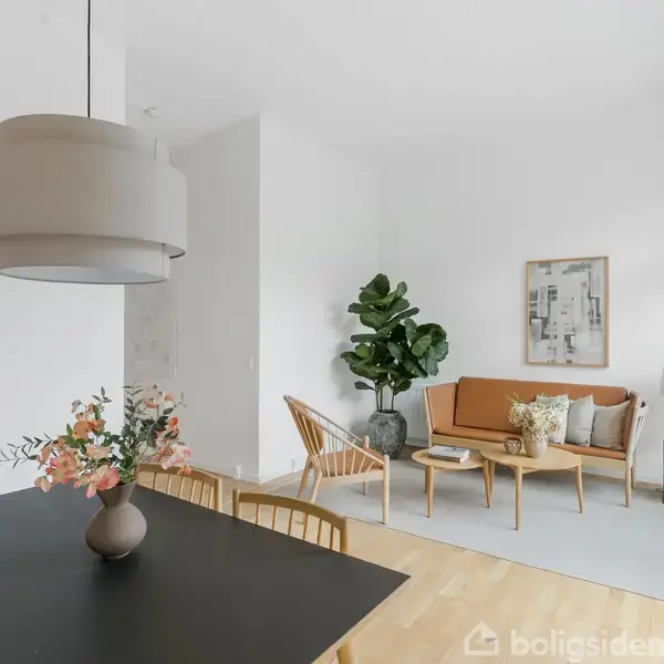 Et stilfuldt, lyst stueområde med en beige sofa, træborde og en gulvlampe. Et spisebord med blomster i forgrunden. Store vinduer giver naturligt lys.