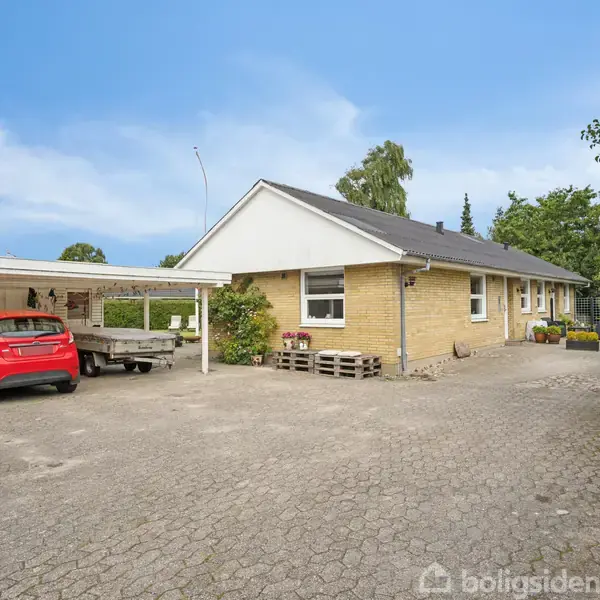 Et gulstenshus med en sort tag ligger i en stor, flisebelagt gårdsplads. En rød bil er parkeret under en åben carport. Grønne buske omkranser området.