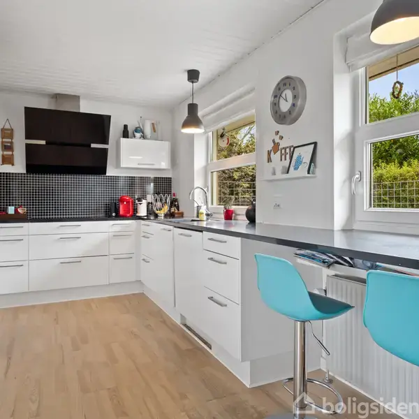 Moderne køkken med hvide skabe og sort flisevæg, har trægulv og to blå barstole ved en høj bordplade. To store vinduer giver udsigt til en grøn have.