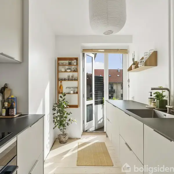 Et smalt køkken med hvide skabe og sorte bordplader. Vinduerne i baggrunden giver naturligt lys, og en åben dør fører til udenfor. Planter og hylder dekorerer rummet.