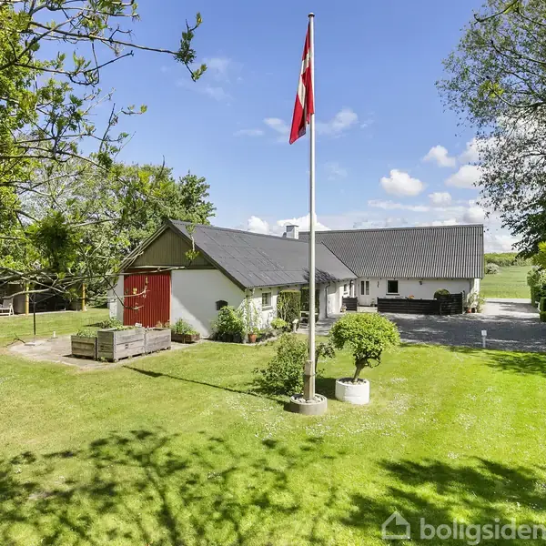 Et rødt og hvidt hus står i en grøn have med en dansk flagstang i forgrunden. Det er omgivet af træer og en blå himmel med få skyer.