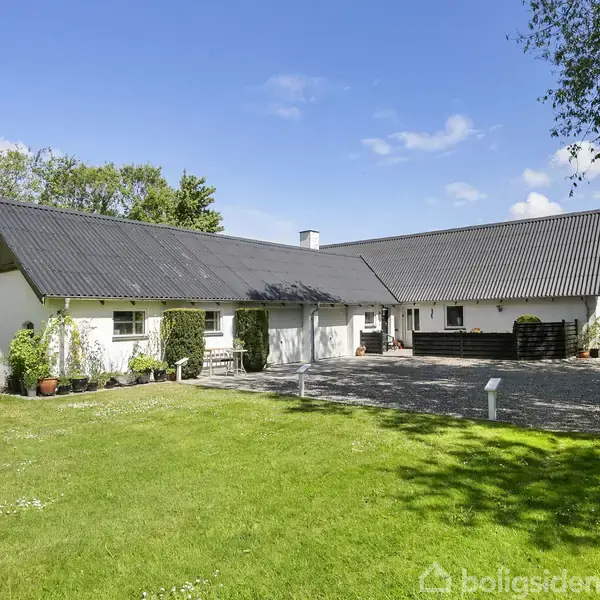 En hvid bygning med sort tag står på græsplæne med træer. Bygningen har rød port og hængende planter. Baggrunden viser en blå himmel med få skyer og omgivelserne er landlige.