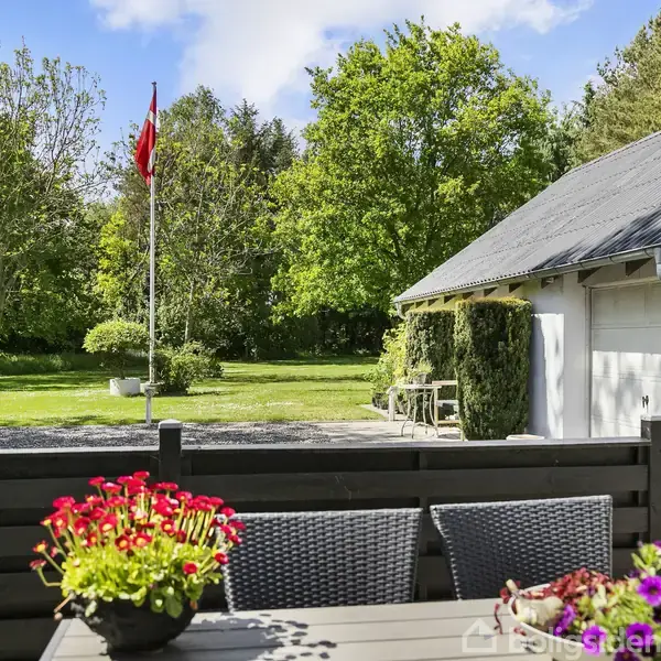 Et udendørs spisebord har blomster i en potte ovenpå. I baggrunden står et hus med garage og en flagstang med det danske flag. Omkring er grøn vegetation og en solrig himmel.