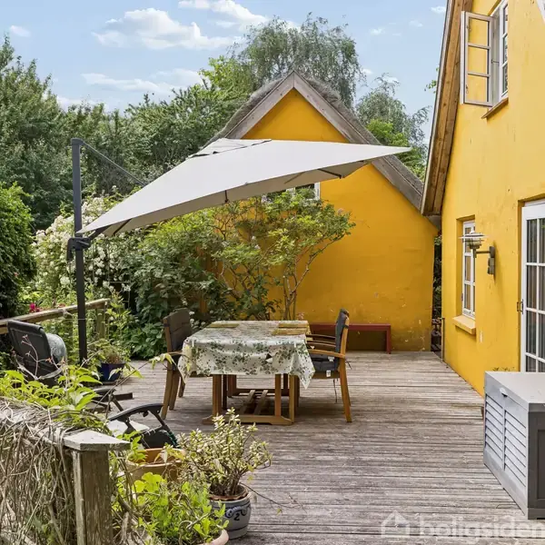 En solrig terrasse med et bord dækket af en dug og stole under en parasol. Den ligger ved siden af et gult hus med stråtag, omgivet af frodig vegetation.