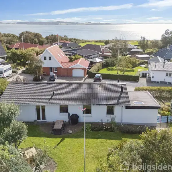 Et enkelt hus med fladt tag står stille i en grøn have omgivet af andre villaer; i baggrunden ses en blå himmel og havudsigt i det fjerne.