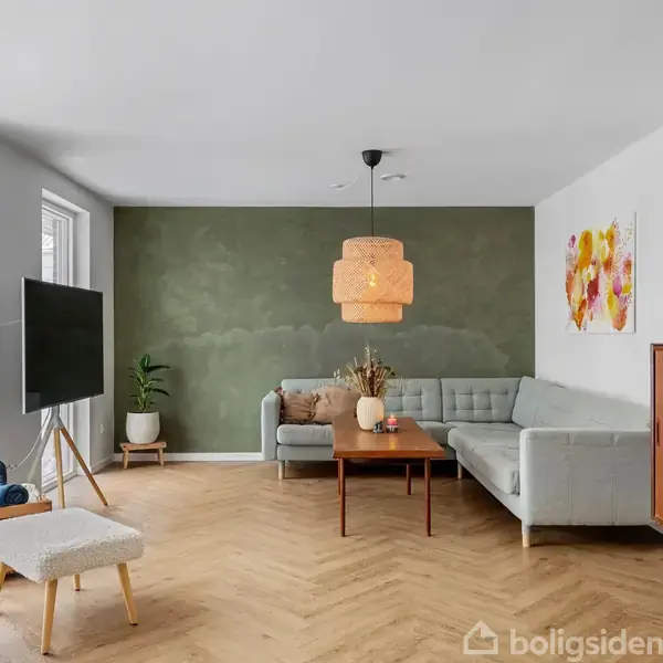 Stue med en grå hjørnesofa og træbord, stående på et parketgulv. En stor lampe hænger over bordet, mens en reol og tv er i rummet. Grøn væg bag sofaen.