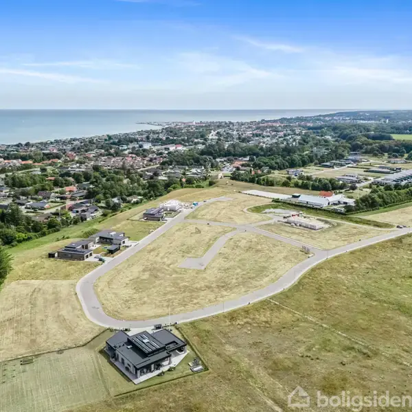 Landområde under udvikling med grusveje snor sig gennem åbne marker. Få bygninger er spredt omkring. I baggrunden ses en by ved kysten mod havet.