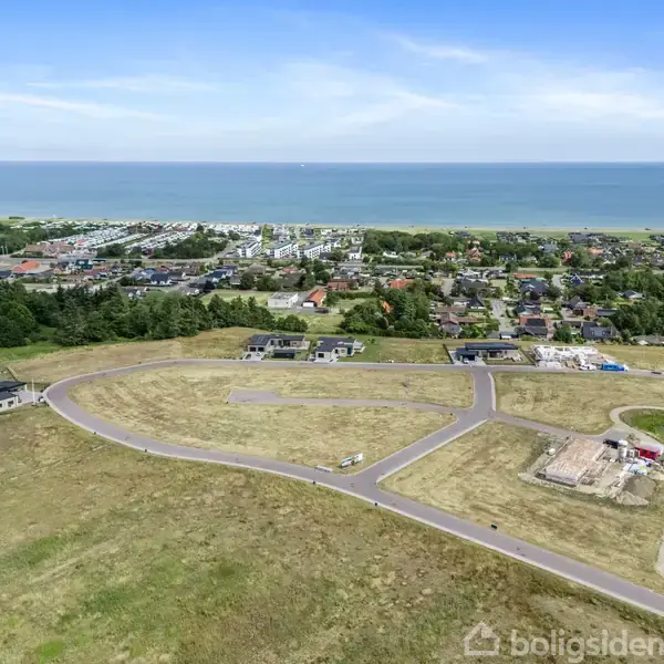 En grusvej med sving og opdelte tomter omgiver en række bygninger under konstruktion. I baggrunden ses en by mod en kystlinje og havet.
