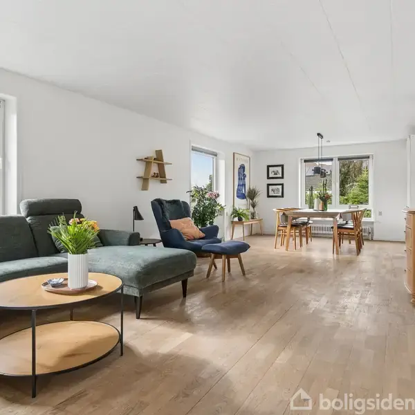 En rummelig stue med en grøn sofa og rundt sofabord. Spisestue med træbord og stole i baggrunden. Store vinduer giver lys, og planter pynter rummet.