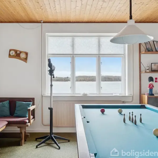 Et billardbord står i et lyst rum med træloft, ved siden af et vindue med udsigt over landskab. En belysningslampe hænger over bordet, og en bænk med puder er i hjørnet.