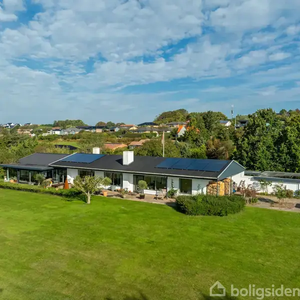 En moderne, langstrakt villa ligger stille omkranset af en frodig have med en stor grøn græsplæne i et landligt boligområde under en blå, skyklar himmel.