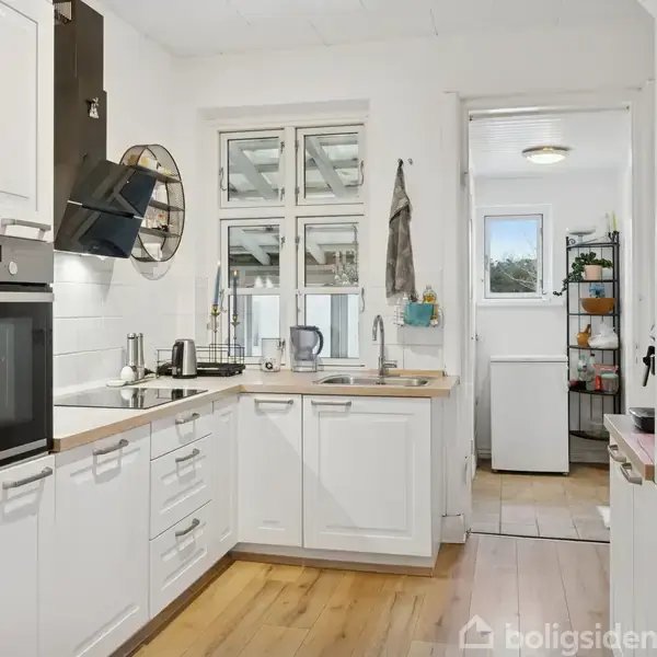 Hvidt køkken med indbyggede skabe og moderne ovn, kogeplade og emhætte, i et lyst rum med trægulv og vinduer over vasken. En dør fører til et tilstødende rum.