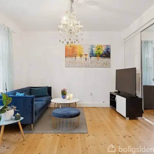 En stue indeholder en blå sofa og sofabord, placeret på et trægulv. En farverig maleri hænger på væggen over sofaen. Et tv står til højre.