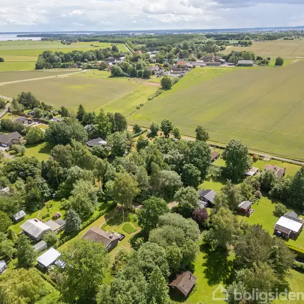 Luftfoto af en landsby med spredte huse omgivet af frodige marker og træer. En landevej strækker sig gennem landskabet, og skyer svæver over horisonten.
