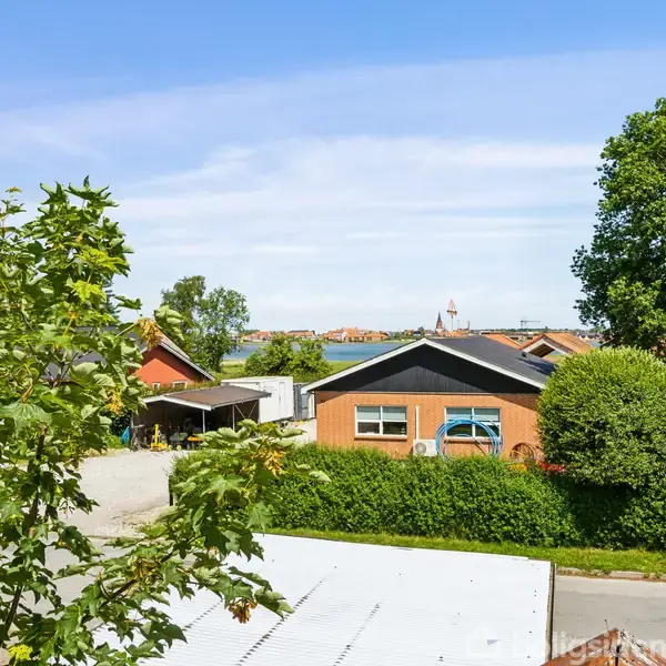 Rækkehuse med røde tage står stille omgivet af grønne træer og buske, mens bygninger vises i baggrunden under en klar blå himmel. En vej løber foran haverne.