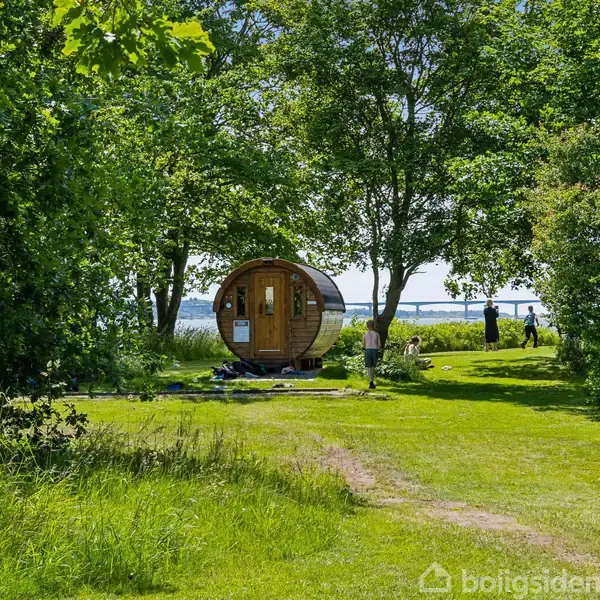En rund, træbeklædt sauna står stille mellem grønne træer med udsigt over en sø eller fjord i baggrunden. Græsset omkring er velplejet, og et par personer opholder sig i nærheden.