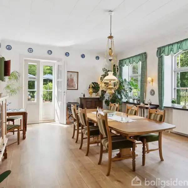 Spisestue med lang træbord omgivet af stole. Et elegant lysekrone hænger over bordet. Grønne planter dekorerer rummet med store vinduer, der giver udsigt til en frodig have.