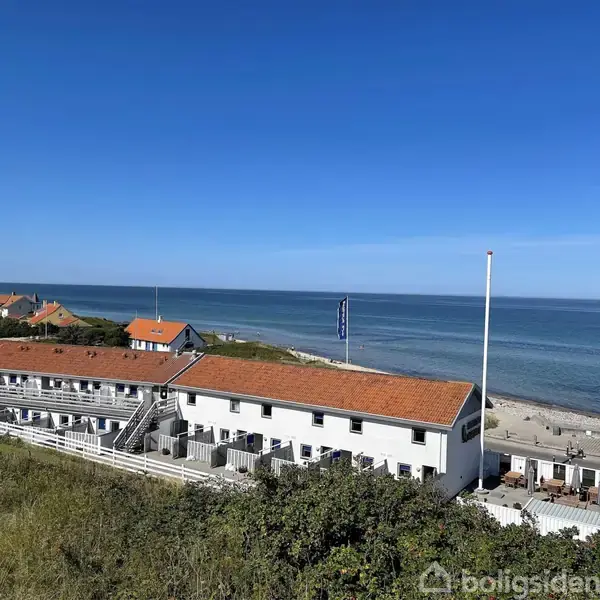 En hvid bygning med rødt tag ligger ved kysten. Bygningen har trapper og terrasser. I baggrunden ses havet, blå himmel og nogle mindre huse.