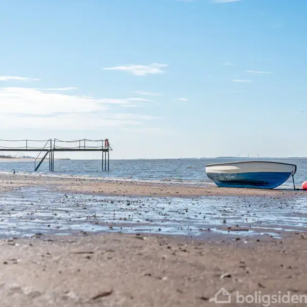 En lille båd ligger på en sandstrand ved lavvande. I baggrunden ses en badebro strække sig ud over vandet under en klar blå himmel.