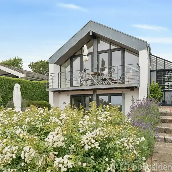 Et moderne hus med glasfacade har en balkon med siddemøbler. Foran huset er en frodig have med blomster og buske. En trappe fører op langs siden.