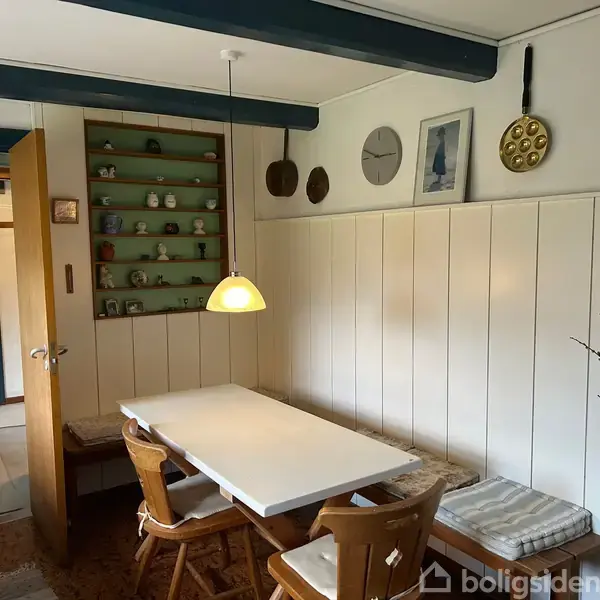 Et hvidt spisebord står i et hyggeligt køkken med to stole og en bænk. På væggen hænger ure og pander, og en hylde viser dekorative genstande.