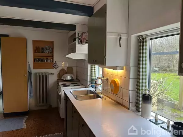 Toilet står ved siden af håndvask i et lille badeværelse. Fliser dækker væggene, et lille vindue lader lys ind. Flasker og håndklæder er placeret nær spejlet over vasken.