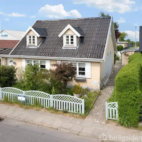 Hus med to kviste på taget står omgivet af en hæk og et hvidt stakit. En sti fører op til døren. Omgivelserne inkluderer en vej og naboer.