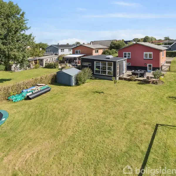 En rød murstensbygning står stille i en stor, grøn have med en trampolin, skur og fodboldmål, omgivet af hegn og under en klar himmel.