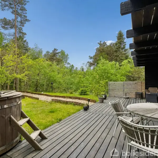 Træterrasse med borde og stole leder mod en træbevokset have; bagerst står en rund, udendørs træbadekar. Skovklædt baggrund under en klar blå himmel.