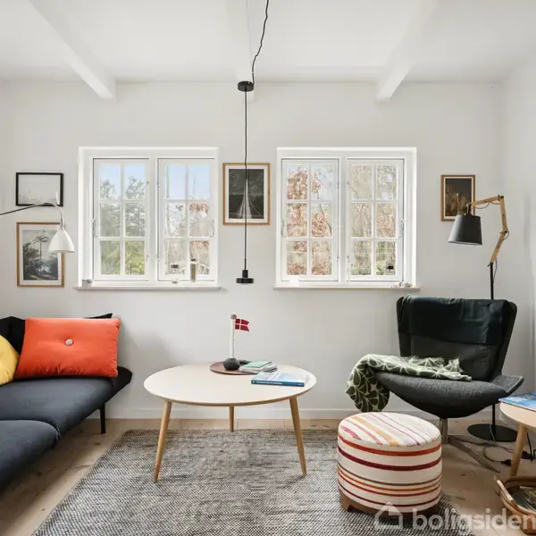 Sofa med farverige puder står ved siden af et rundt sofabord med bøger, i et lyst rum med store vinduer. En lænestol og en stribet puf står tæt ved et TV.