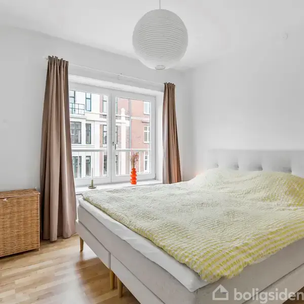 Soveværelse med en dobbeltseng dækket af et gulnet tæppe, placeret ved et stort vindue med beige gardiner. Ved siden af står en kurvevasketøjskurv og en lille natbord.