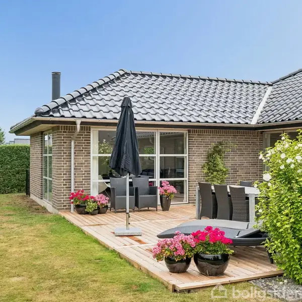 Et moderne hus med gråt tegltag og store vinduespartier. På terrassen står et sort parasol og havemøbler omgivet af pink blomsterkrukker. Græsplæne og hække omkranser området.