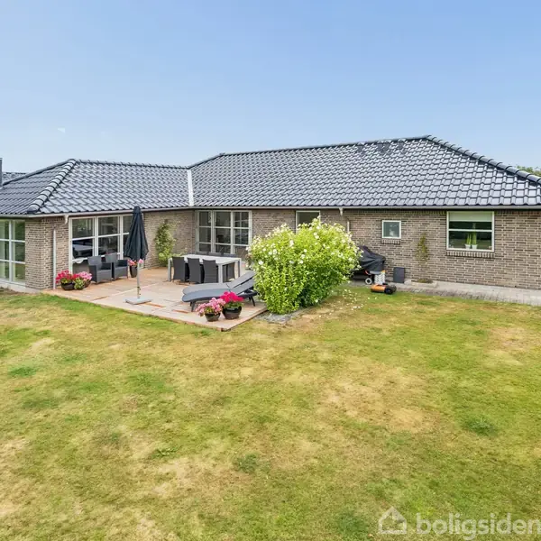Huset står stille på en velplejet græsplæne med terrasse, hvor et bord og stole er placeret. Murstensfacaden har store vinduer, og der er få buske rundt om huset.