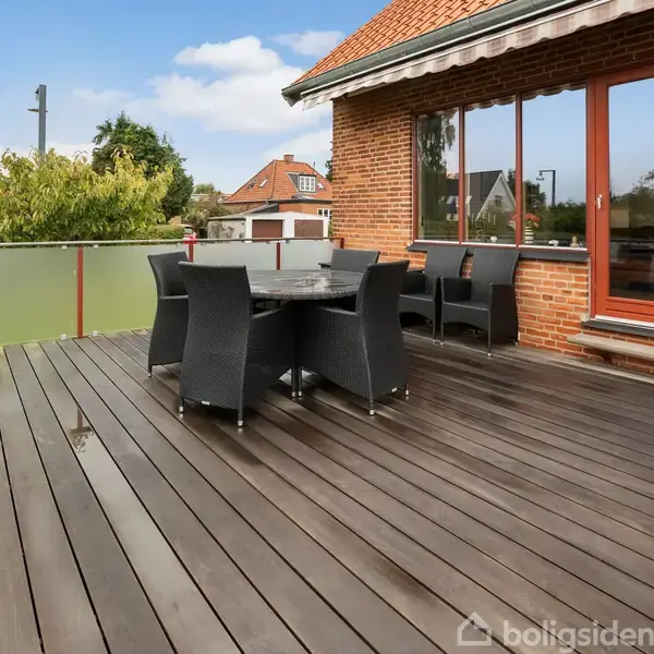 Terrassen har et sæt sorte kurvemøbler placeret på træplanker. Den er omgivet af et gelænder med matte glasplader. Den rødmurede bygning i baggrunden har store vinduer og en dør.