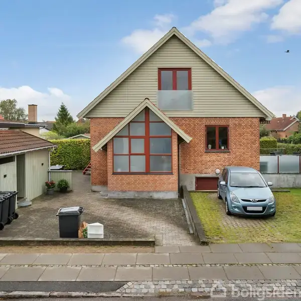 Et rødt murstenshus med stort, trekantet vindue står i en forstadshave. En bil er parkeret på græsplænen ved siden af. Bagved er en garage med affaldsbeholdere.