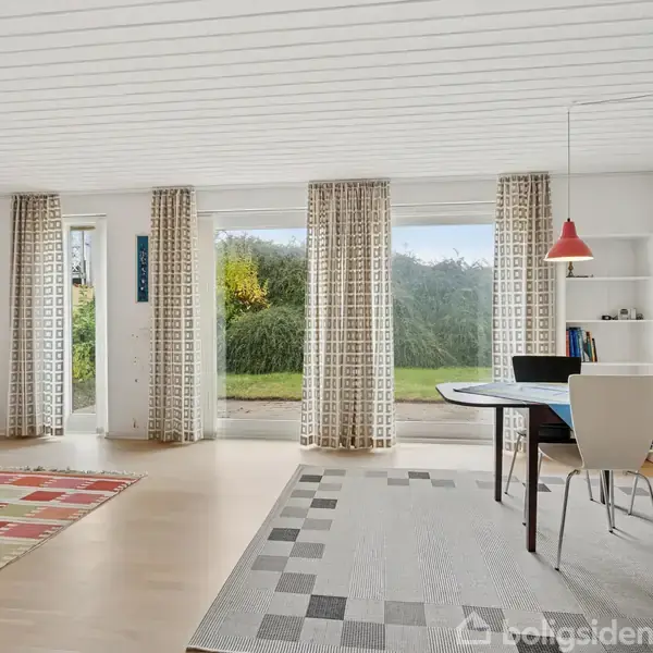 Et lyst stueområde med et rundt spisebord og stole placeret på et geometrisk mønstret tæppe. Store vinduer med gardiner viser en grøn have udenfor.