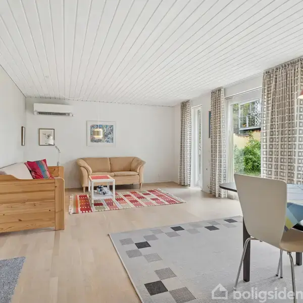 Stue med en lys sofa og to stole står på et farverigt tæppe. Et spisebord med stole er ved siden af store vinduer. Lysekrone hænger over bordet.