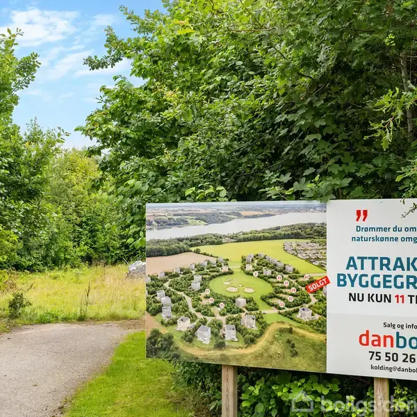 Et skilt reklamerer for byggegrunde med teksten: "Drømmer du om at bo i naturskønne omgivelser? ATTRAKTIVE BYGGEGRUNDE NU KUN 11 TILBAGE. Salg og info: danbolig 75 50 26 00 kolding@danbolig.dk." Det står ved en skovsti omgivet af træer.