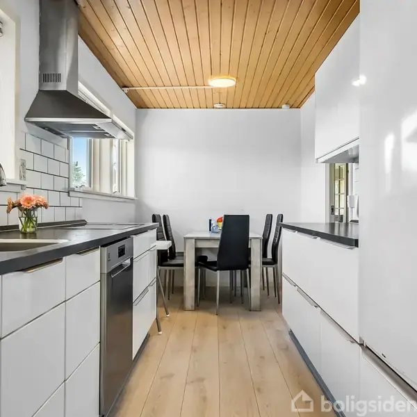 Et moderne køkken med hvide skabe og sort bordplade. En vask og opvaskemaskine på venstre side. Et spisebord med fire stole i baggrunden; træloft og lyse trægulve.