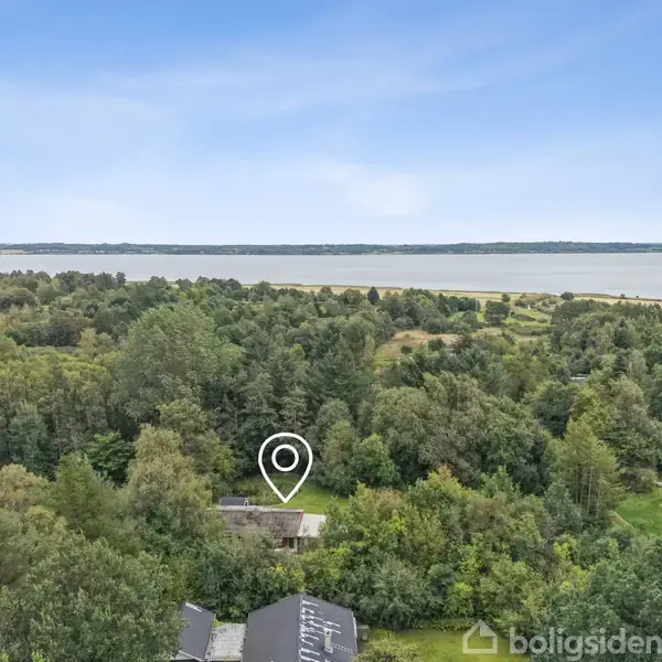 Hus markeret med en nål ligger omgivet af tæt skov og frodig vegetation. Baggrunden viser en bred flod eller sø med en klar, blå himmel.