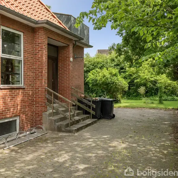 En rød murstensbygning med trapper, hvor tre affaldsspande står ved siden af. Omkring bygningen er der grønne buske og træer, som danner en frodig haveindgang.