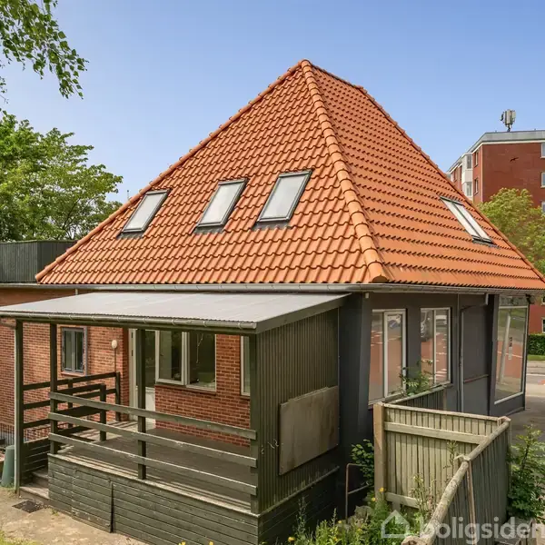 Hus med rødt tegltag og flere kvistvinduer står stille ved vejen. Omkring huset er der træer og en flisebelagt gårdhave. I baggrunden ses en lejlighedsbygning.