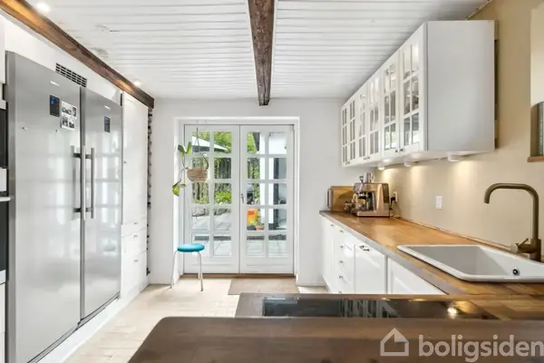 Et køkken med hvide skabe og mørkt træbord. Til venstre er store stålkøleskabe, og i midten ses en dør med en stol foran, der fører ud til en terrasse.